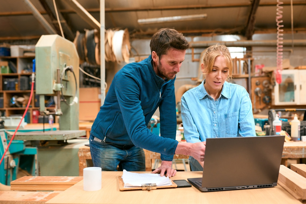 Two people in a carpentry shop look at a laptop