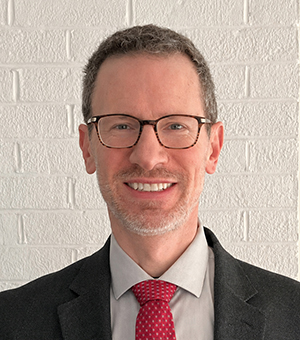 Norman Jones headshot in a black suit, gray shirt and scarlet tie