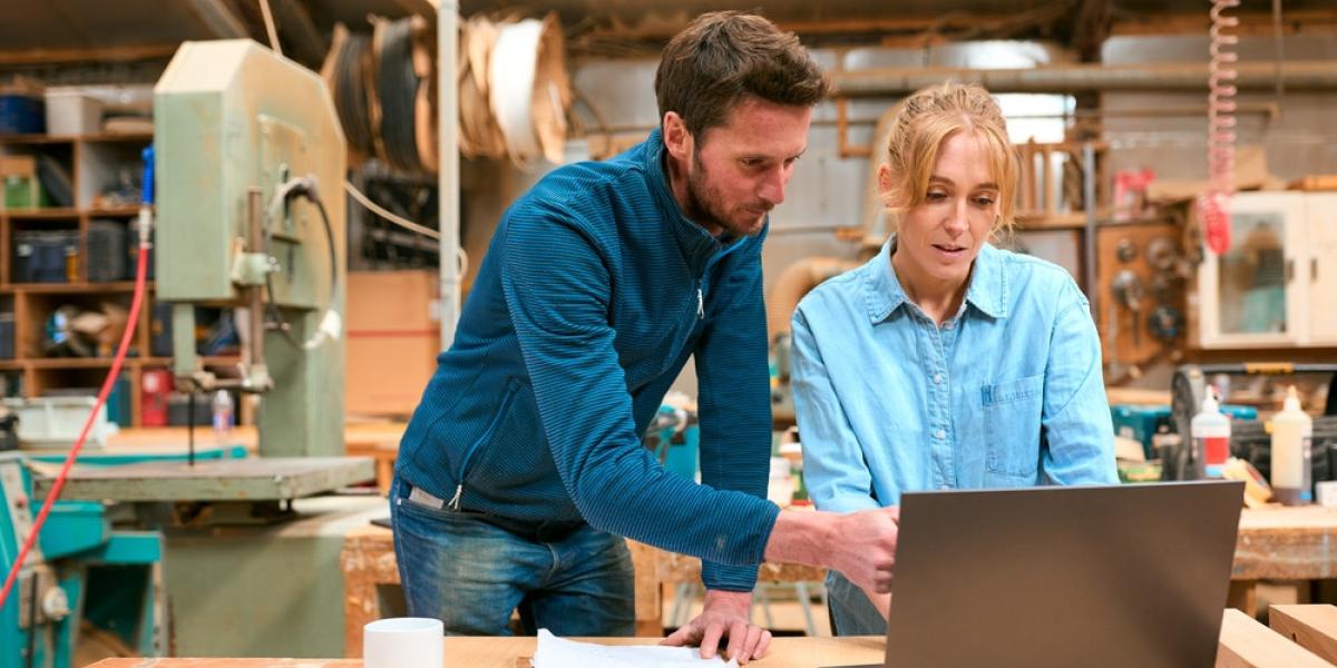 Two people in a carpentry shop look at a laptop