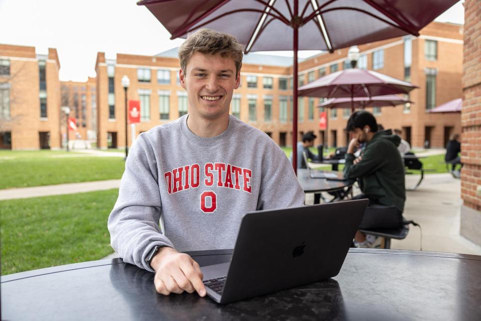 A smiling student with a laptop sitting outside on campus.