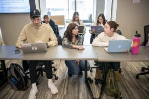 Faculty member kneels between two students using laptops. 